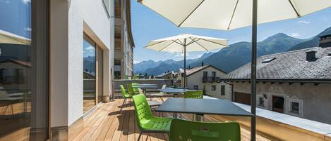 Eine Terrasse mit Holzboden, grünen Stühlen und einem Tisch unter einem großen Sonnenschirm mit Blick auf eine gebirgige Landschaft.