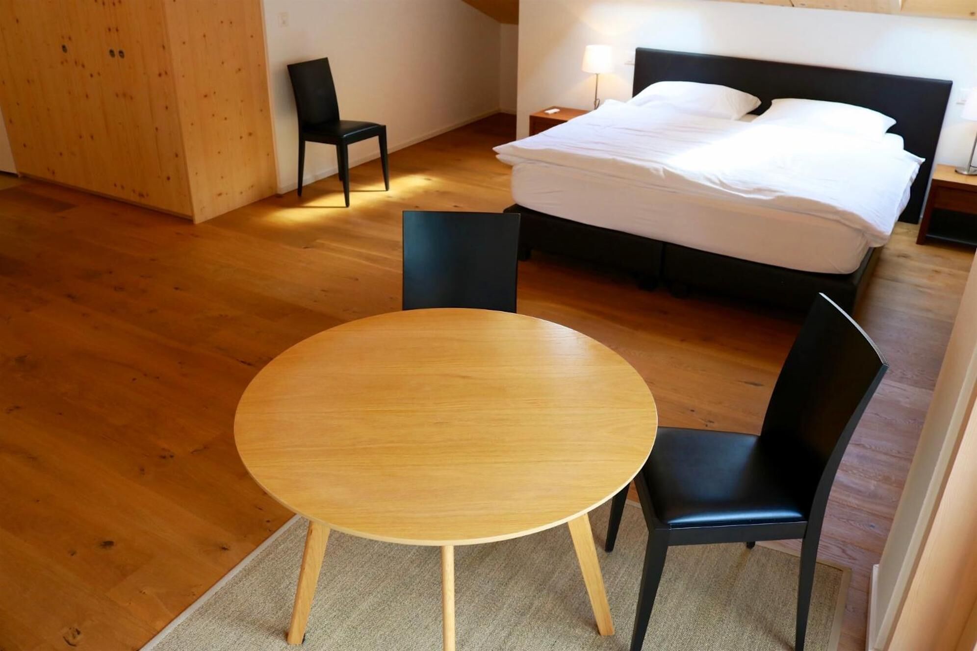 A hotel room with a wooden floor, a round table with chairs, a bed with white linens, and a nightstand with a lamp.