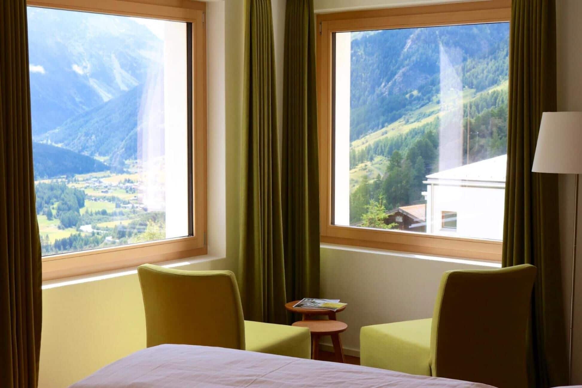 Une chambre d’hôtel avec deux grandes fenêtres donnant sur les montagnes, un lit, deux chaises et une petite table avec un livre.