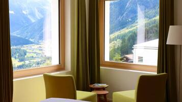 Une chambre d’hôtel avec deux grandes fenêtres donnant sur les montagnes, un lit, deux chaises et une petite table avec un livre.