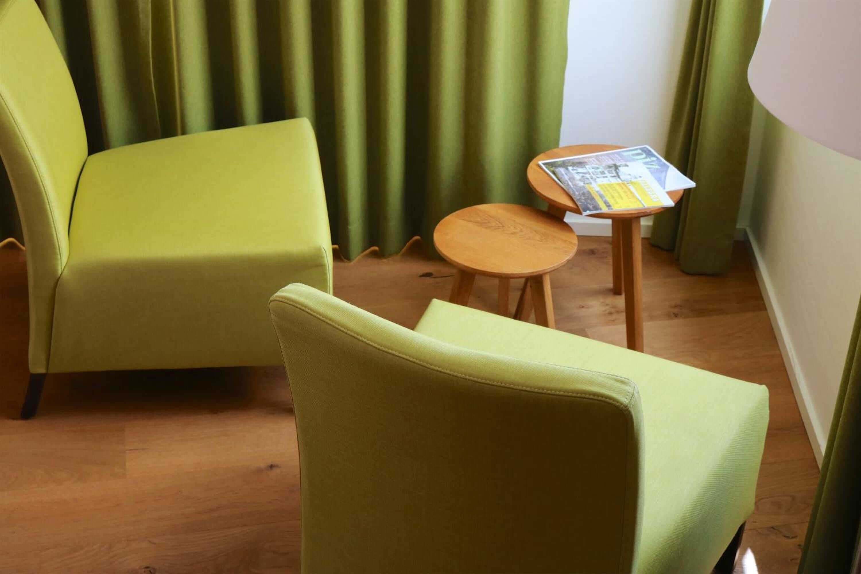 Um quarto com uma poltrona verde, uma pequena mesa de madeira com uma revista e uma cortina verde.