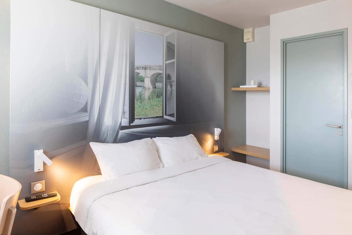 Une chambre d’hôtel avec un lit, des lampes de chevet, une table de nuit, une fenêtre donnant sur un pont et une porte.