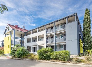 Un edificio de varios pisos con balcones, una entrada visible y vegetación circundante.