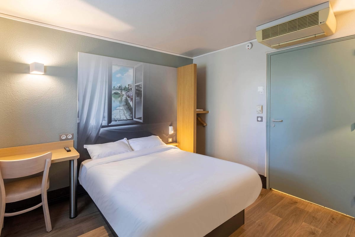 Une chambre d’hôtel avec un lit, un bureau, une chaise, un tableau et une porte.