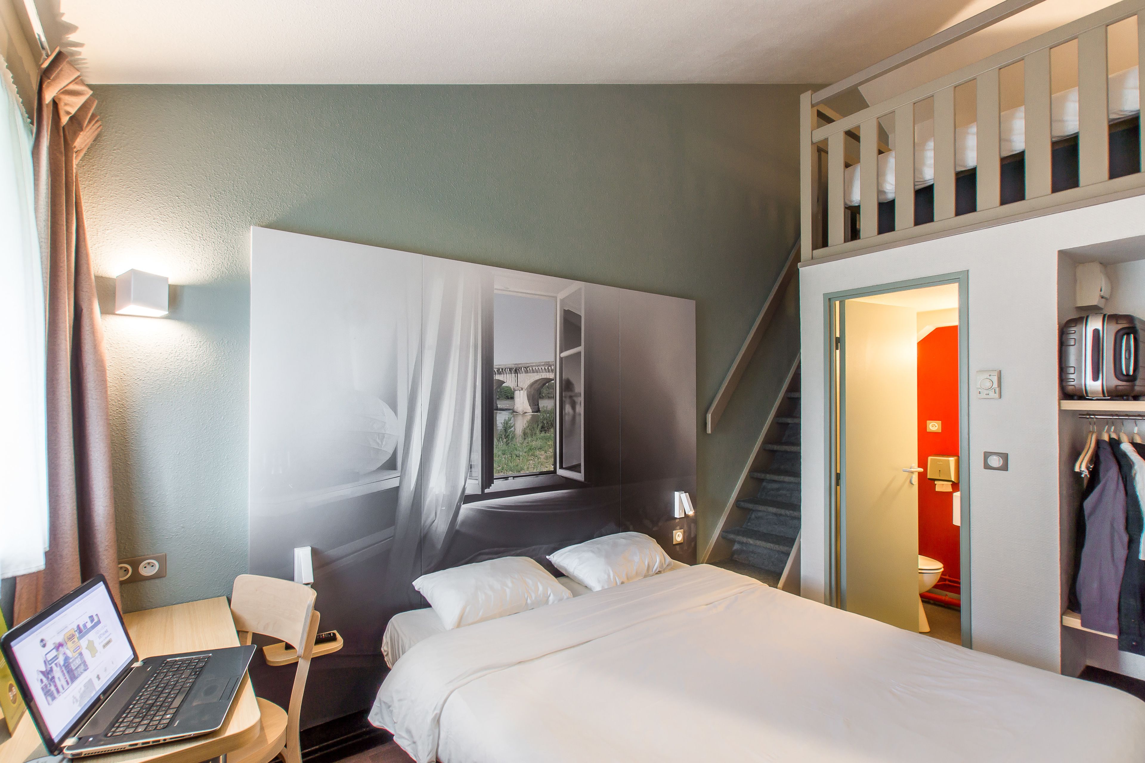 Habitación de hotel con cama, escritorio con computadora portátil, escalera y vistas al exterior.