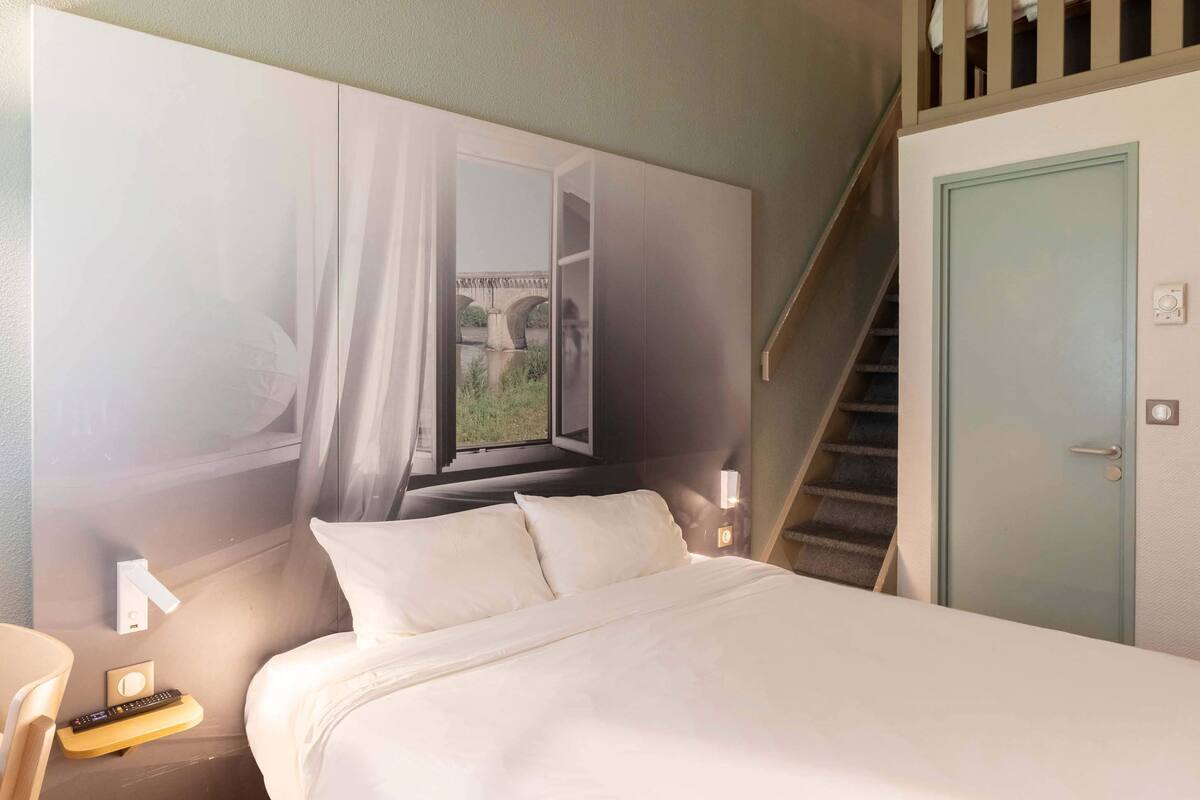 Une chambre d’hôtel comprenant un lit, une table de chevet, une télécommande, une lampe fixée au mur et une porte avec un interrupteur.
