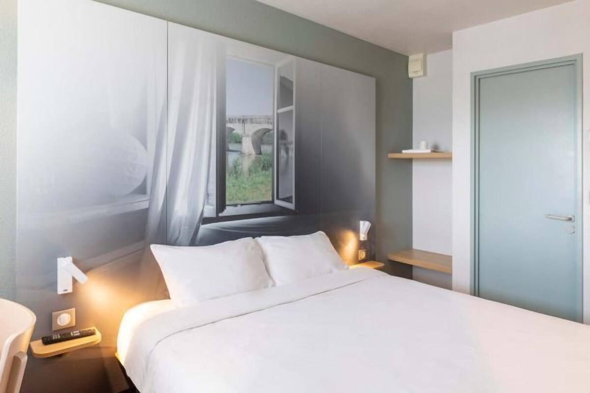 A hotel room with a bed, bedside tables, a nightstand, a window with curtains, a door, and a shelf.