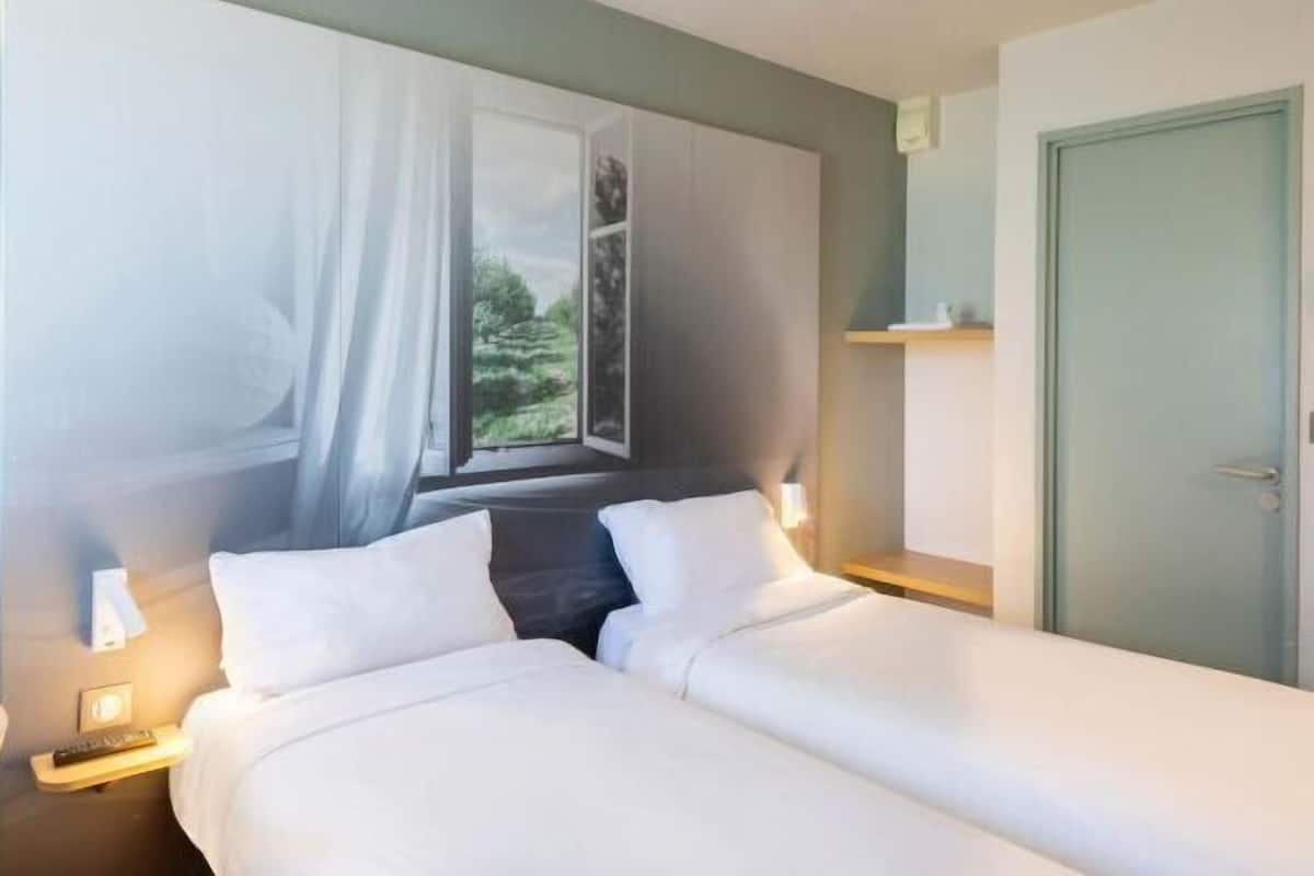 Une chambre d’hôtel avec deux lits, une fenêtre donnant sur un paysage agréable, une porte et une table de chevet avec une lampe.