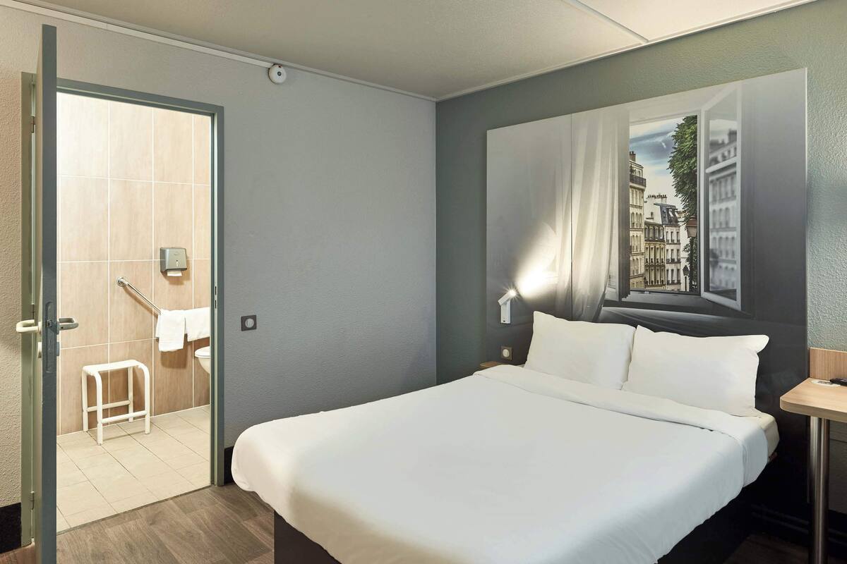 A hotel room with a large bed, a nightstand, a wall-mounted lamp, and a painting of a cityscape.