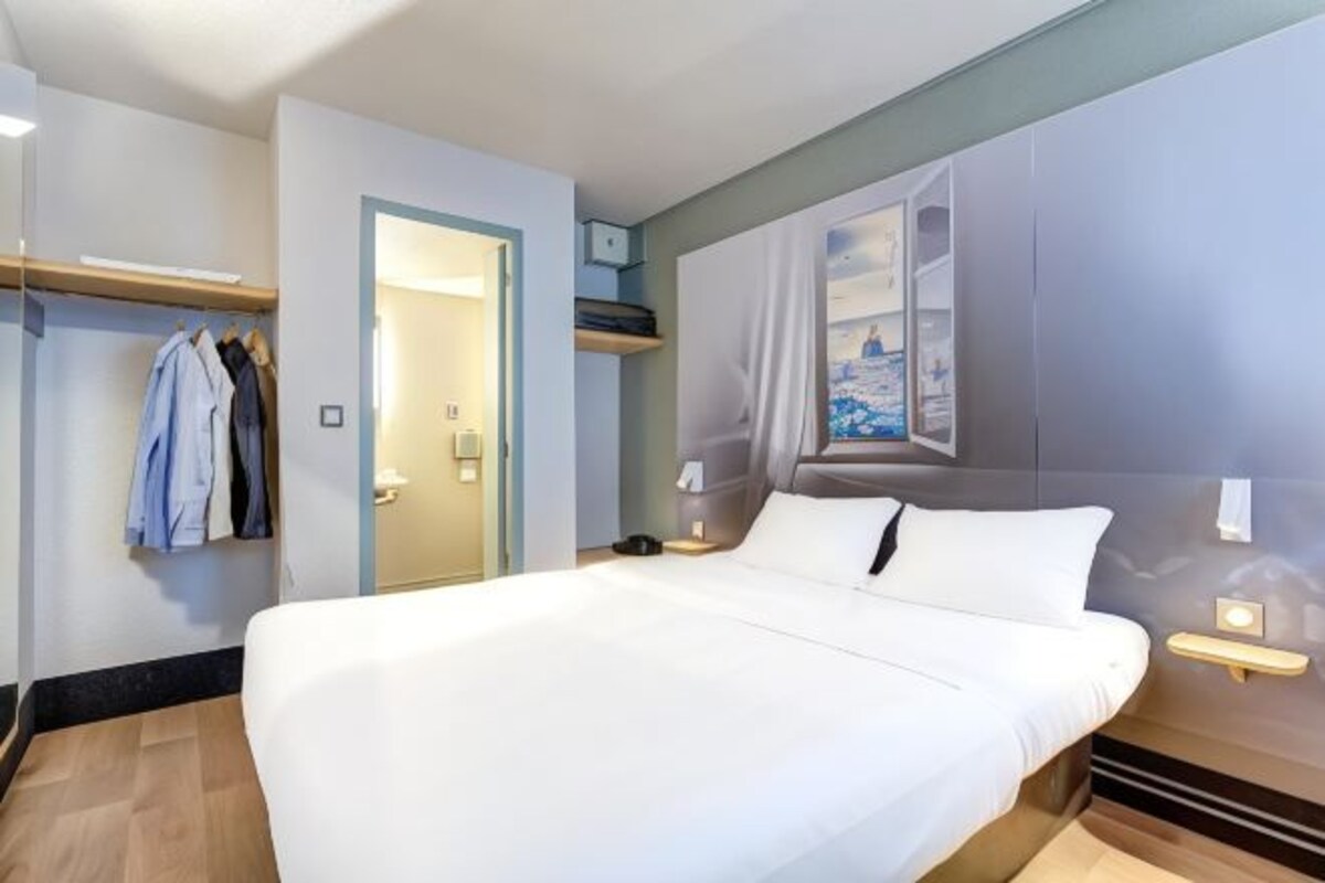 A hotel room with a bed, a coat rack, a door leading to a bathroom, and a painting on the wall.