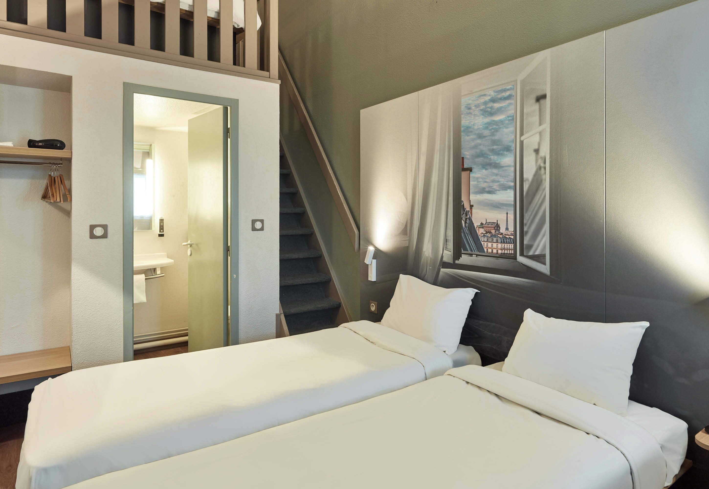 A hotel room with a bed, a bathroom, a staircase, and a cityscape painting on the wall.