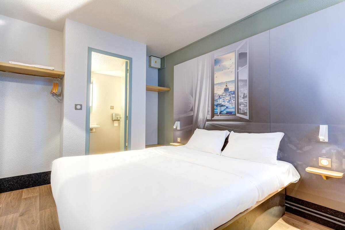 A modern hotel room with a large bed, a wooden floor, a wall-mounted shelf, and a door leading to another room.