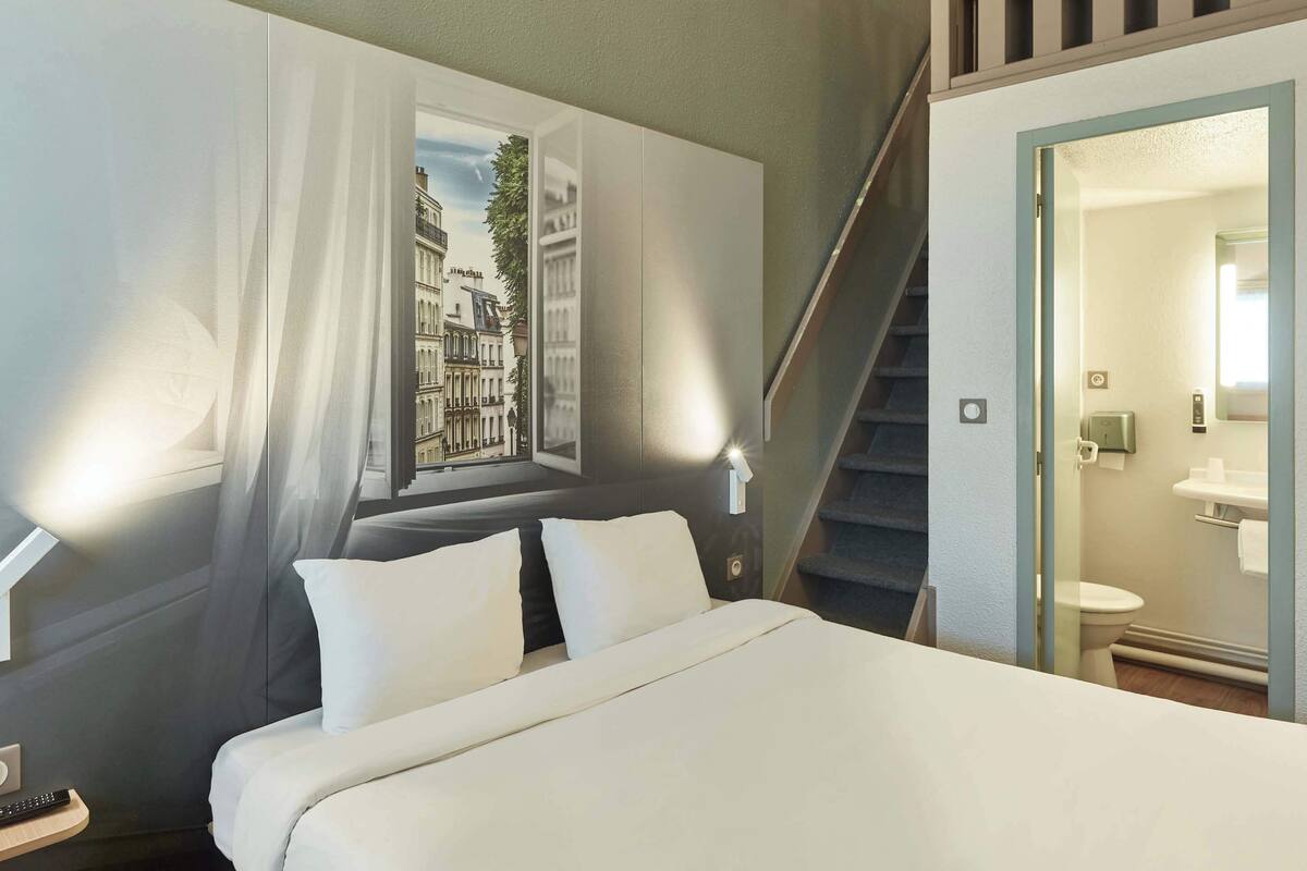 A hotel room with a bed, a nightstand, a remote control, a wall-mounted lamp, a window with curtains, a staircase, and a bathroom visible through an open door.