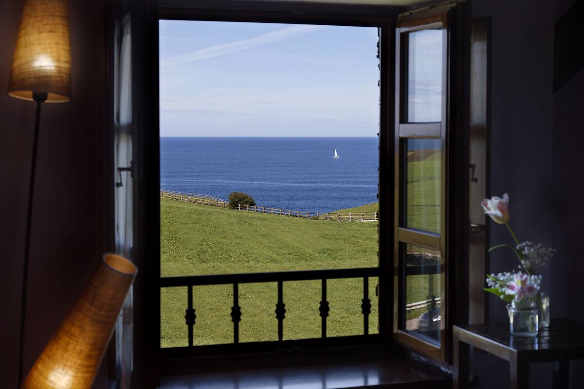 A room with a view of the sea, a balcony with a railing, and a vase with flowers on a table.