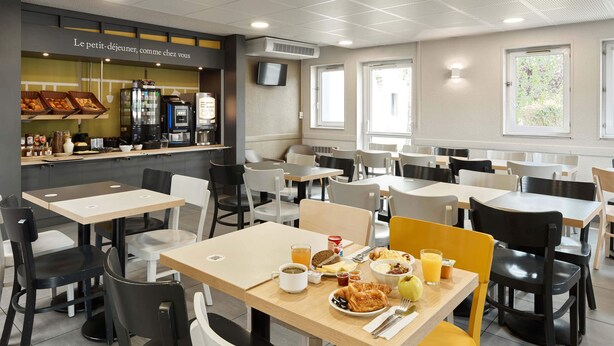 A café with tables and chairs, a counter with food items, and a television on the wall. - B&B HOTEL Paris Nord Villepinte (Villepinte)