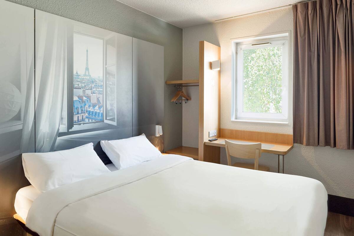 Een hotelkamer met een groot bed, een bureau en een stoel. Er is een raam met uitzicht op een stadsgezicht en een gordijn.