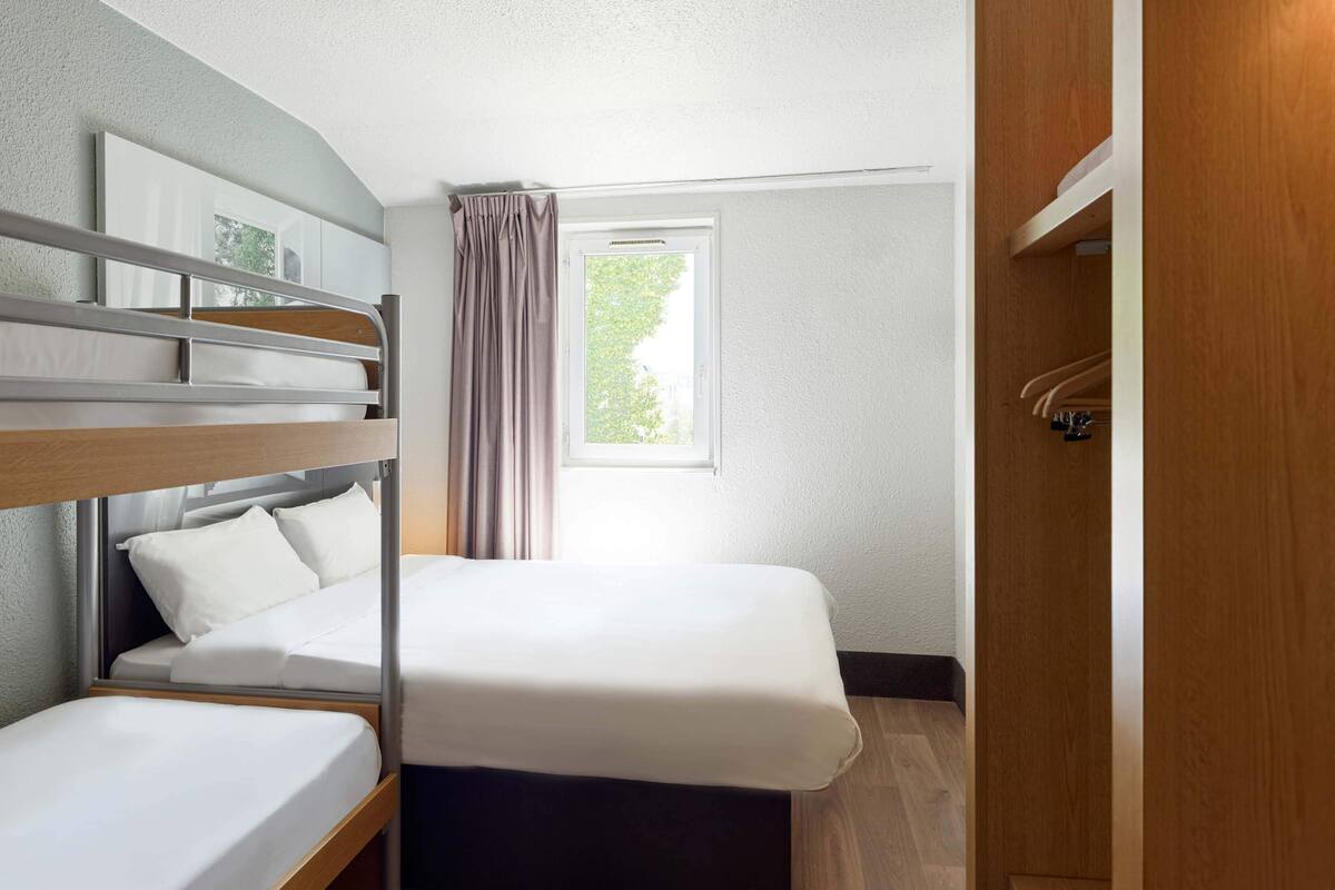 Een compacte hotelkamer met een stapelbed, een enkel bed, een raam met gordijnen en een houten garderobe.