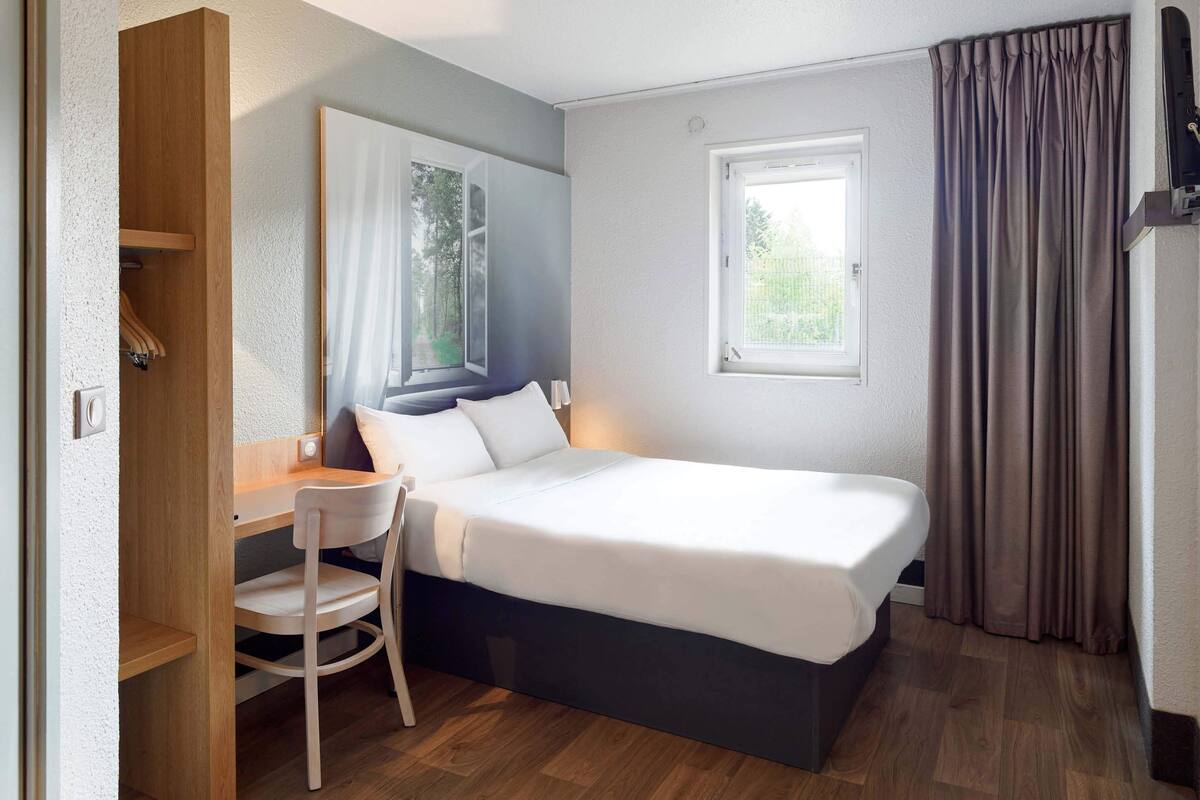 Een compacte kamer met een bed, een bureau en een stoel. Er is een raam met gordijnen en uitzicht op groen buiten.