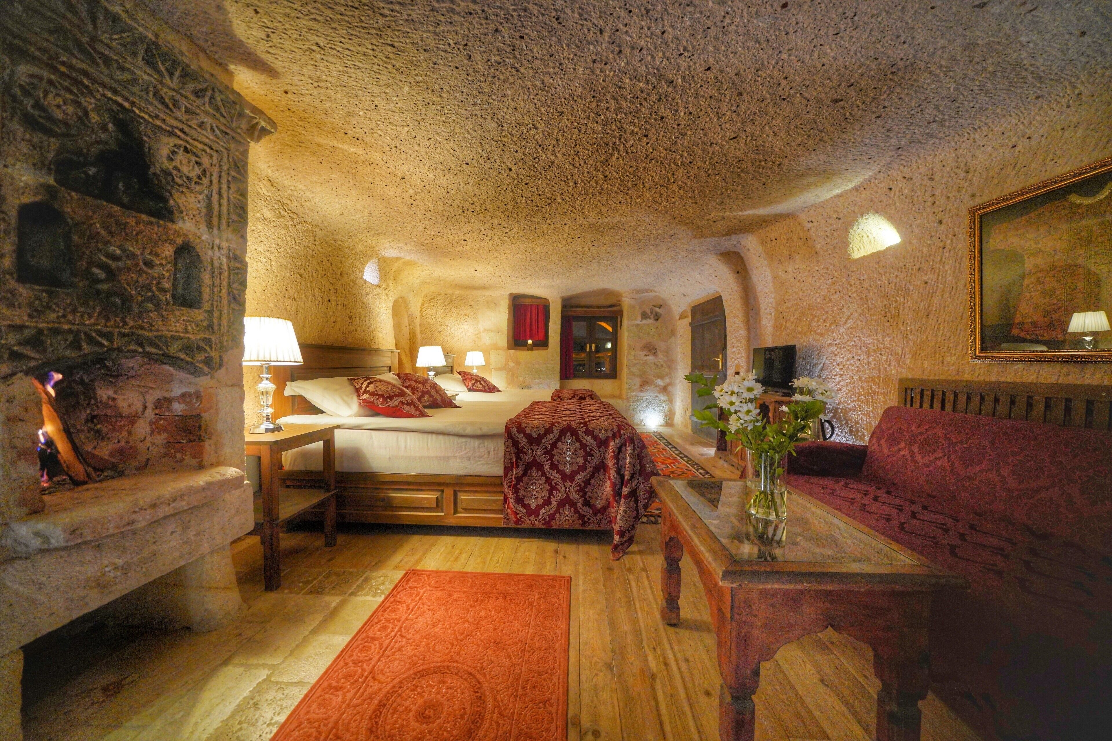 A cozy room with a stone wall, a bed with red bedding, a wooden bench, a red sofa, and a small table with a vase of flowers.
