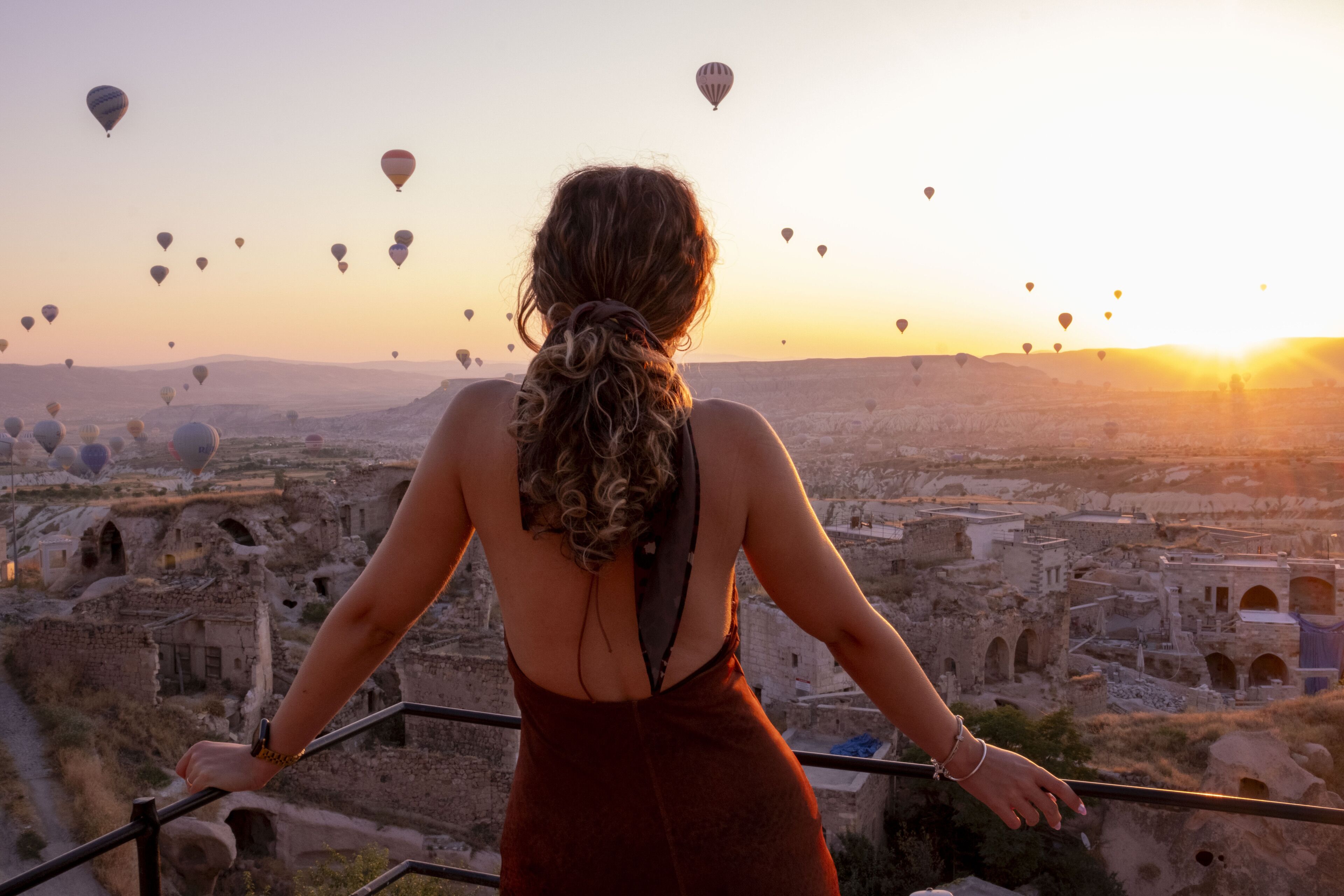 A woman standing on a balcony overlooking a historic cityscape with numerous hot air balloons in the sky.