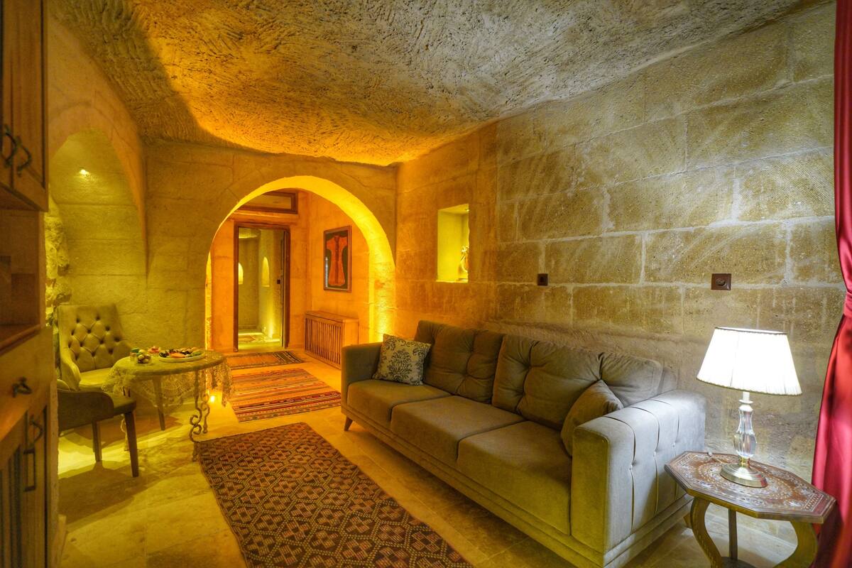 Un cuarto acogedor con un sofá, un sillón y una pequeña mesa con una lámpara. Hay una alfombra estampada en el suelo y una pared de piedra.