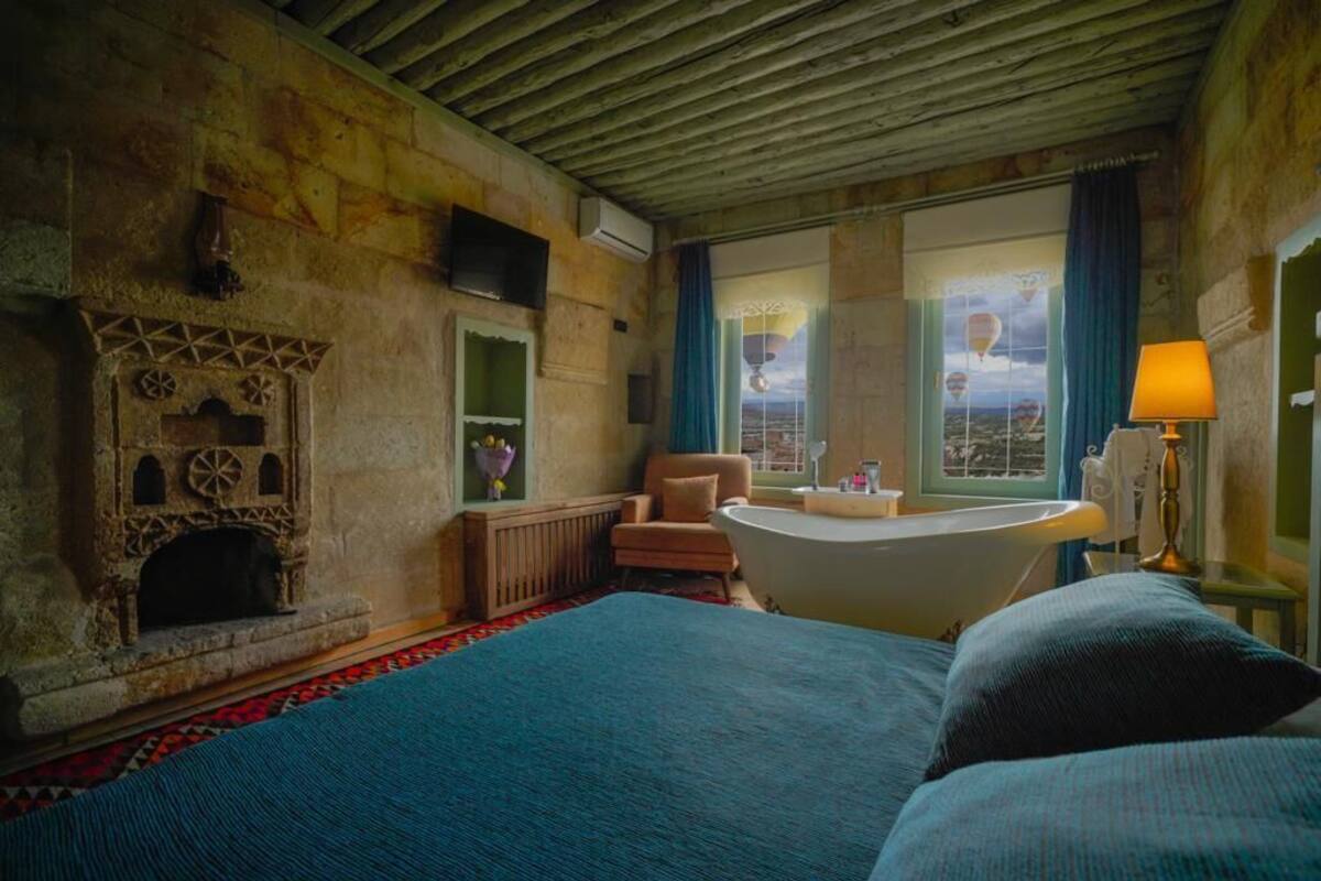 Una habitación con chimenea de piedra, una bañera, una cama grande, una silla y vistas a globos aerostáticos a través de una ventana.