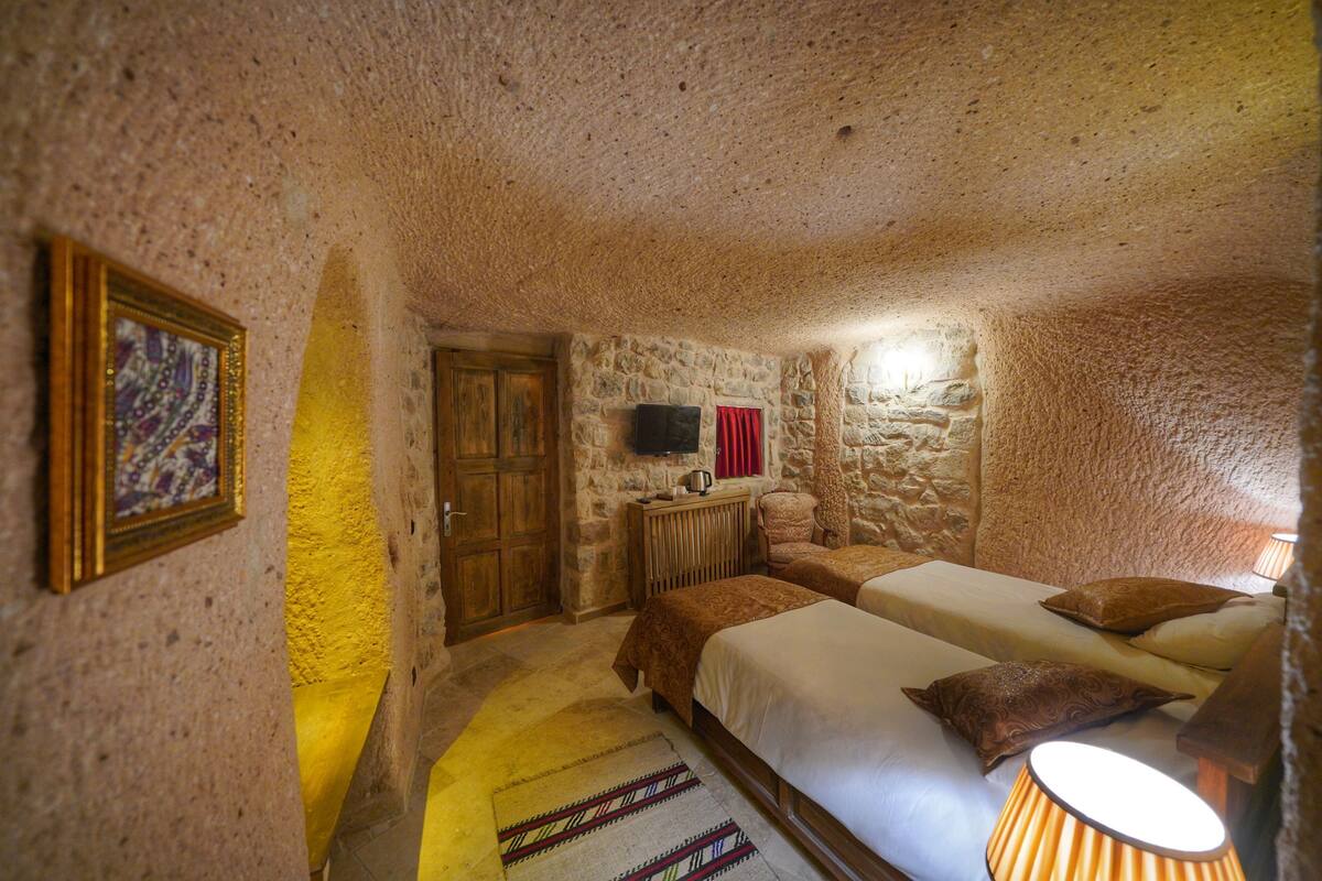 Una habitación rústica con paredes de piedra, una puerta de madera, una cama con colcha estampada, una mesita con lámpara y un televisor montado en la pared.