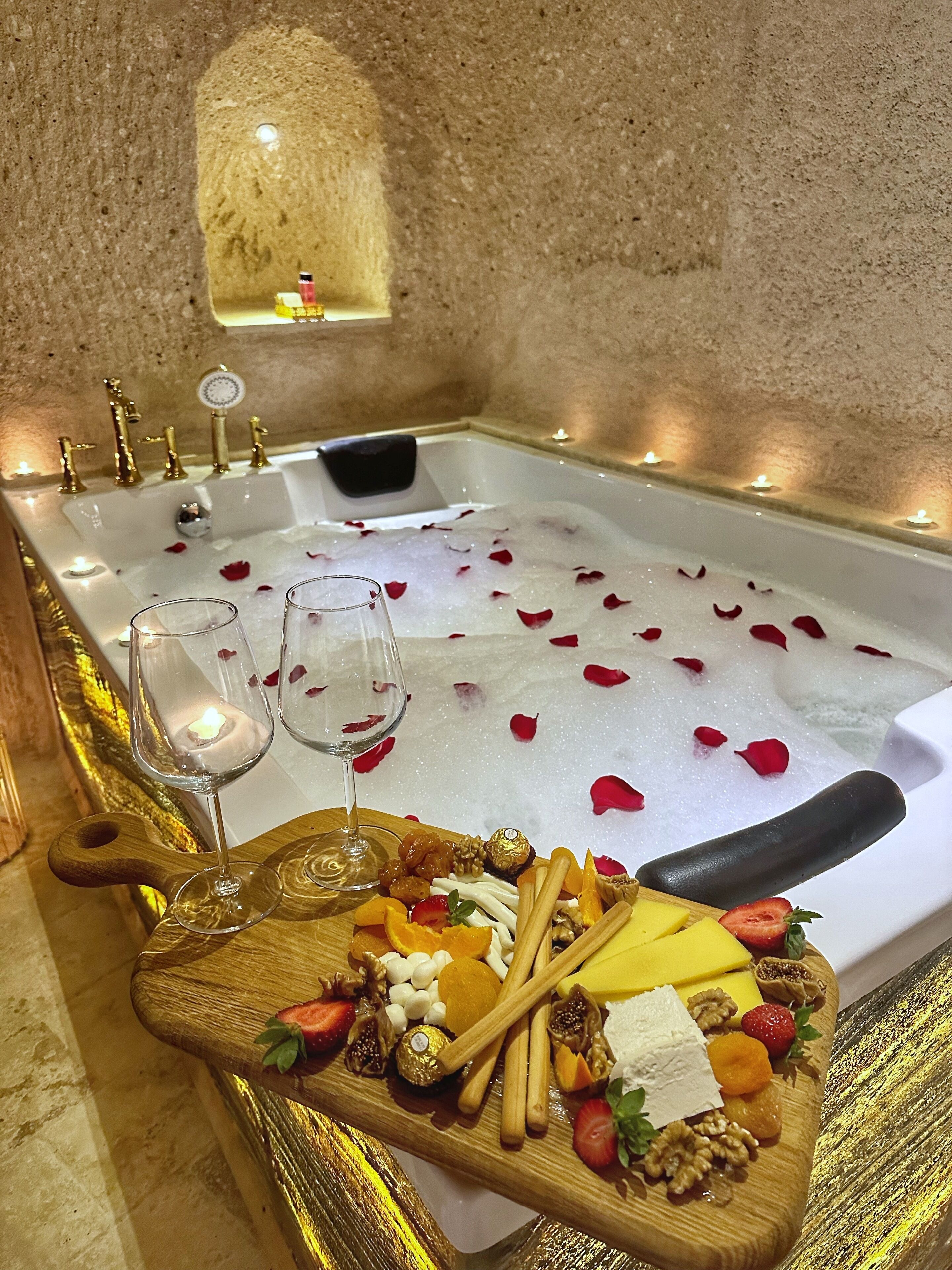 A bathtub filled with bubbles and rose petals, with two glasses and a tray of fruits and nuts beside it.