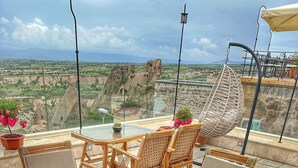 Mesa de vidro redonda com cadeiras de vime e um vaso de planta em um terraço com vista para uma paisagem rochosa.