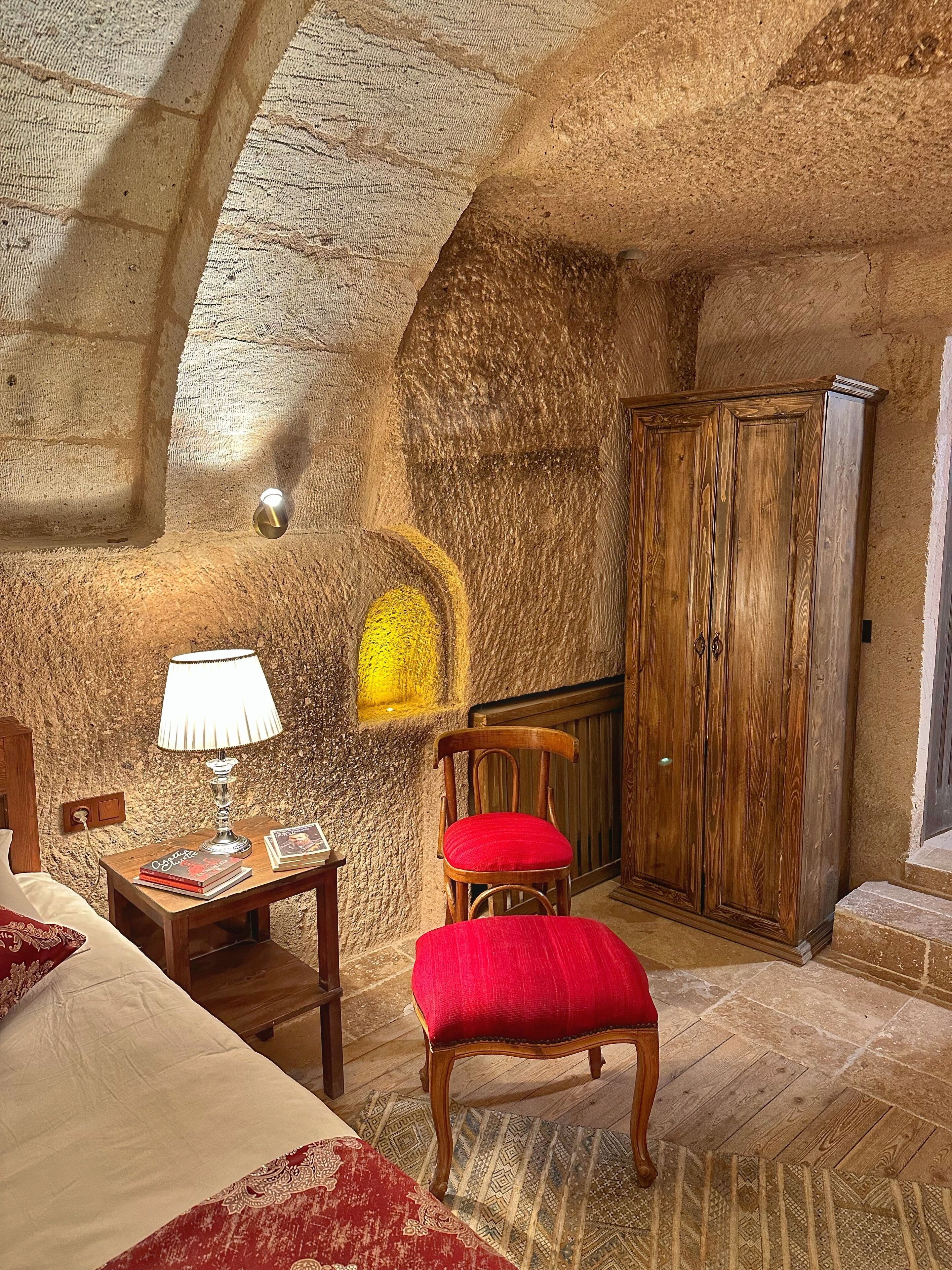 A rustic room with a wooden chair, a wooden cabinet, a lamp, and a bed with a red and white patterned bedspread.