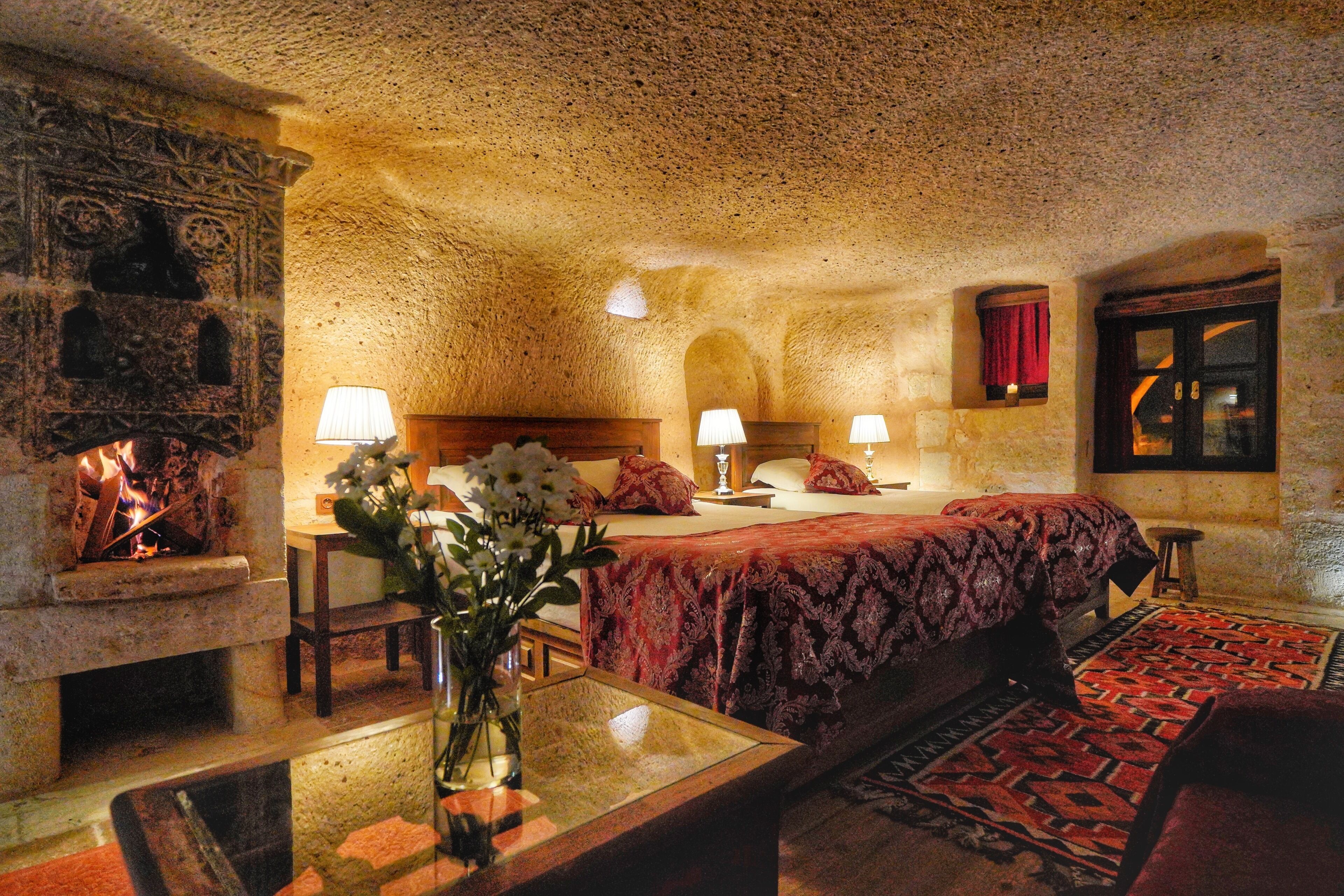 A cozy room with a stone fireplace, a bed with red bedding, a wooden nightstand, and a vase with flowers.