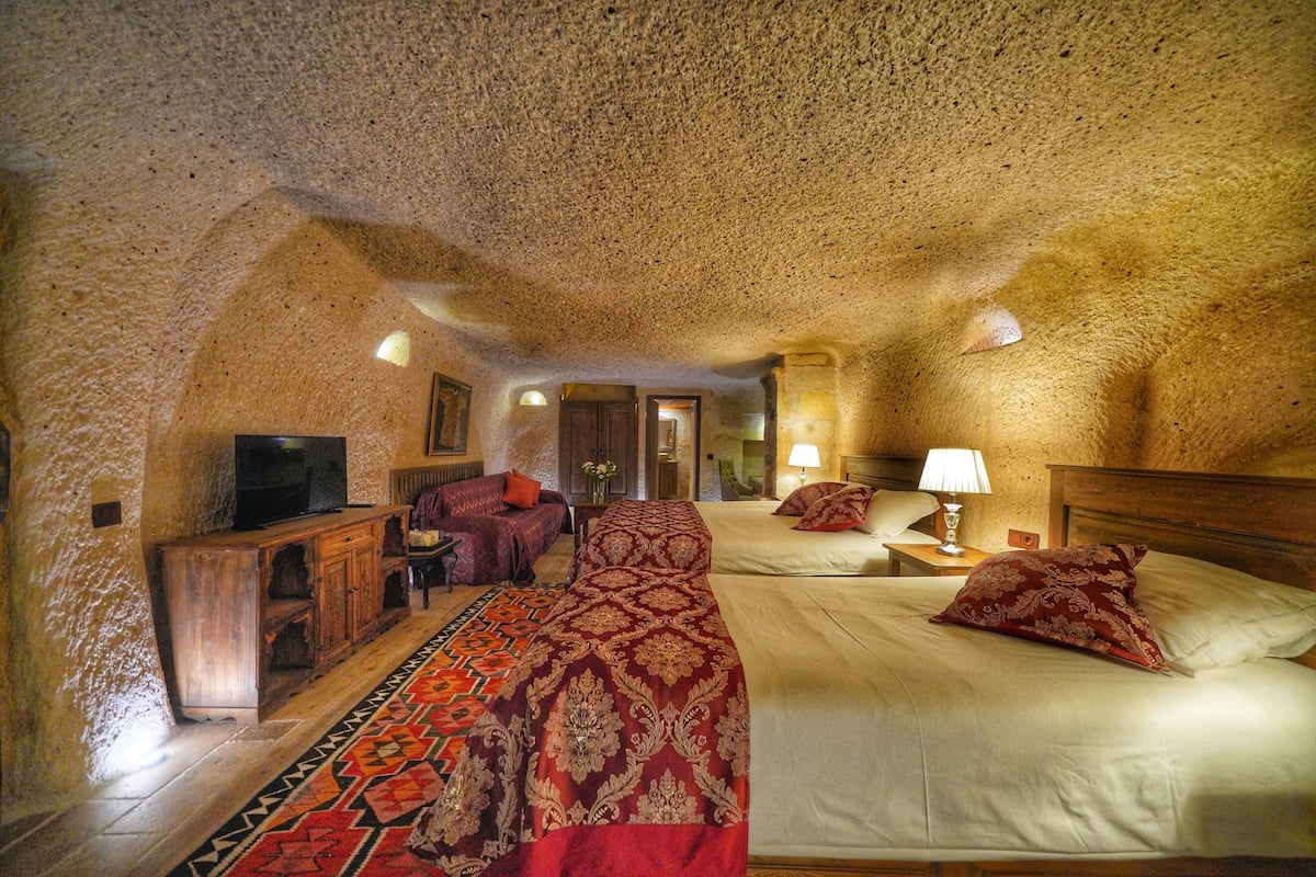 Una habitación con un techo alto y texturizado, una cama con colcha estampada, un televisor sobre un mueble de madera y una mesita con lámpara.