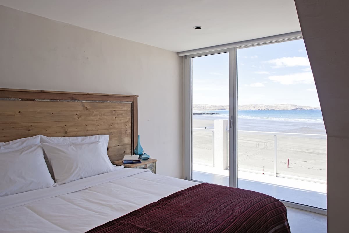 A bedroom with a large bed, a wooden headboard, a nightstand with a vase, and a view of the beach through a sliding glass door.