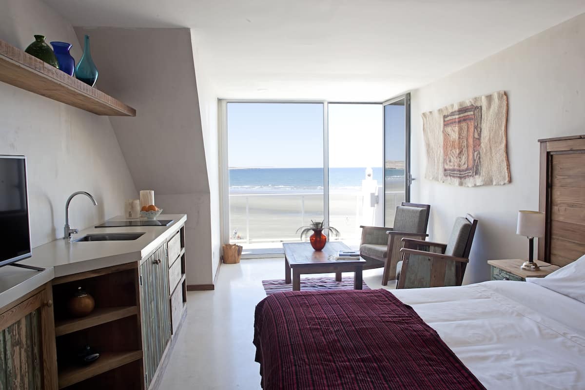 A bedroom with a large bed, a desk, a chair, a TV, and a view of the beach.