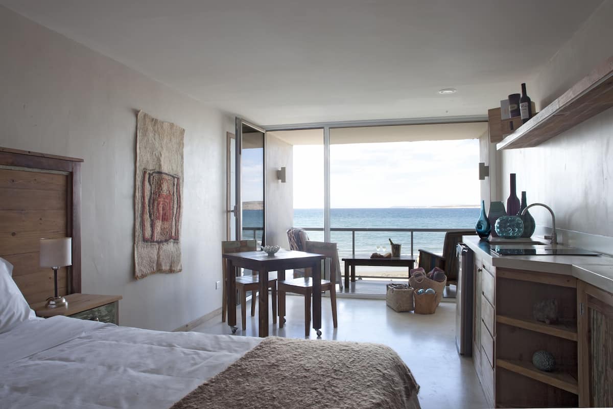 A bedroom with a large bed, a wooden nightstand, a table with chairs, a view of the sea through a sliding door, and a kitchenette area.