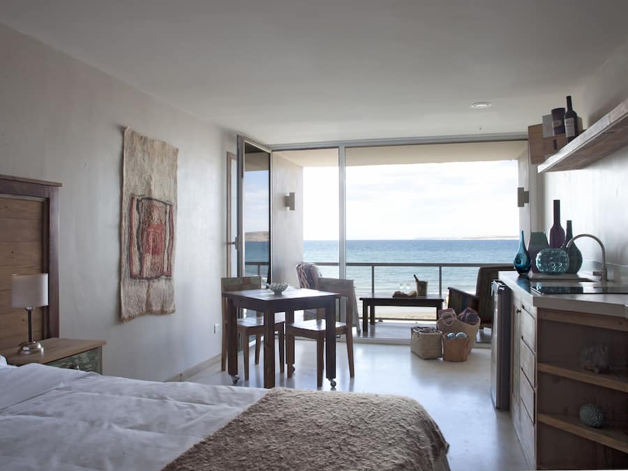Une chambre dotée d’un grand lit, d’une table de chevet en bois, d’une table avec des chaises, d’une vue sur la mer accessible par une porte coulissante, et d’un coin cuisine.