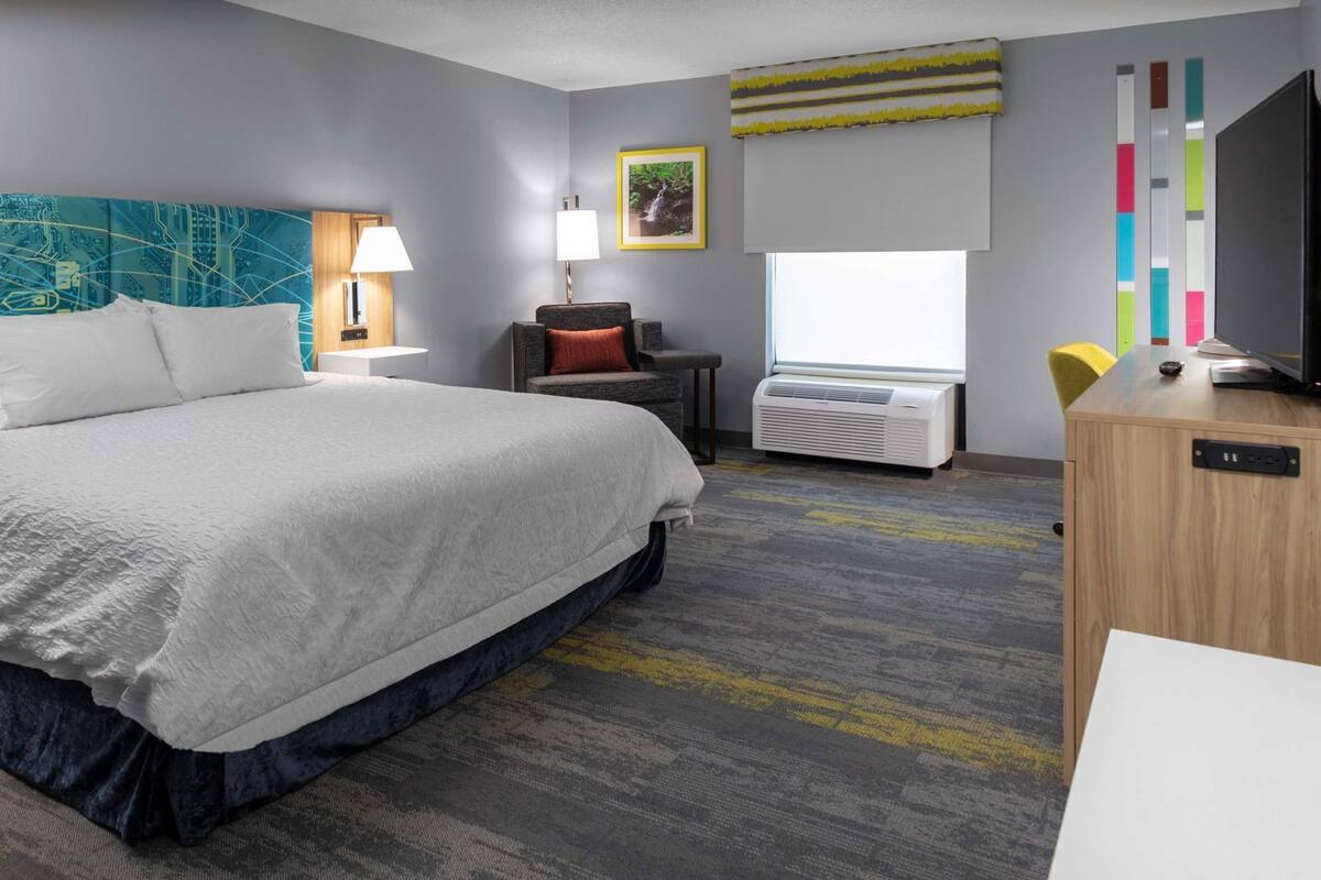A hotel room with a large bed, a desk with a TV, a window, and a wall with a colorful artwork.