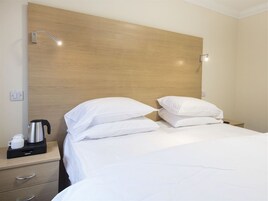 Habitación de hotel con cabecera de madera, cama con ropa blanca, mesita de noche con hervidor y taza, y lámpara de pared.