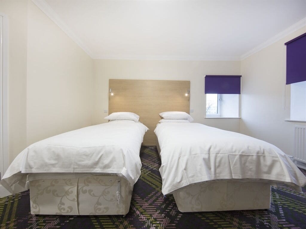 Une chambre d’hôtel avec deux lits simples, une tête de lit, une fenêtre avec un store violet et une moquette à motifs.