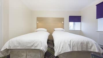 Une chambre d’hôtel avec deux lits simples, une tête de lit, une fenêtre avec un store violet et une moquette à motifs.