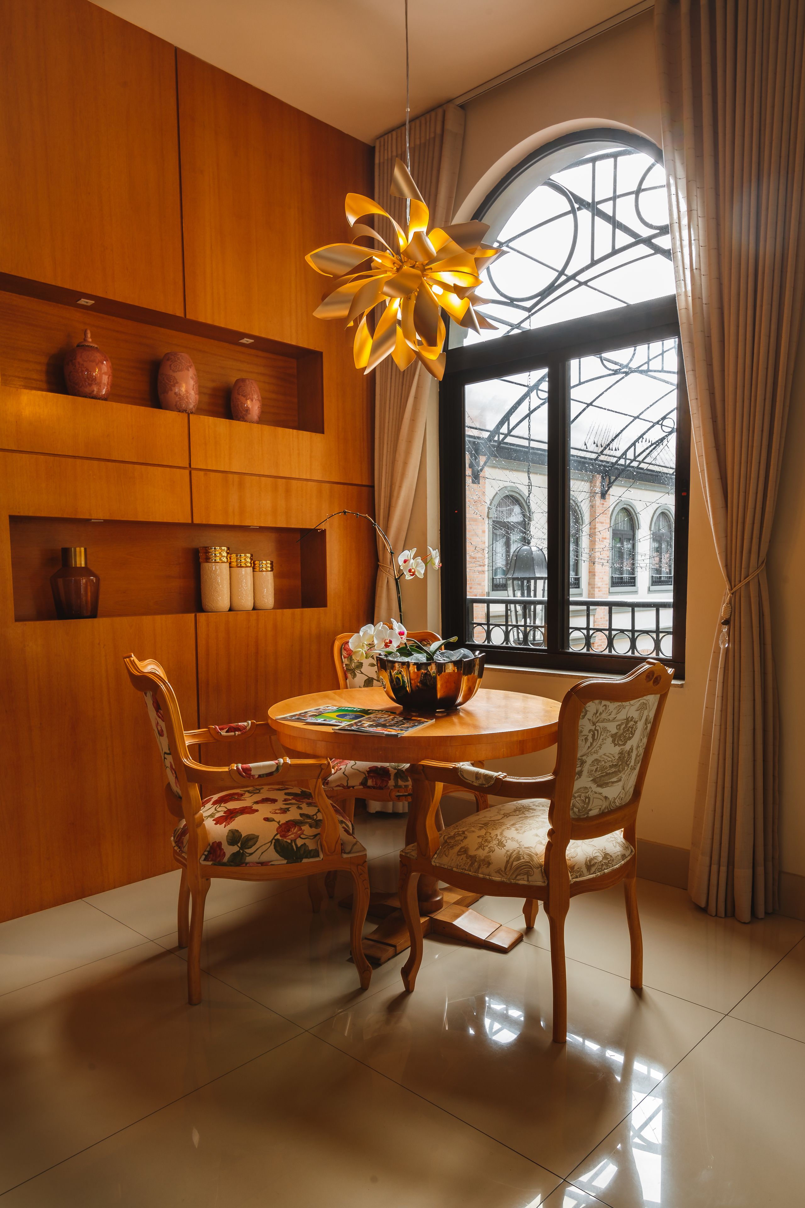A dining area with a wooden table, floral chairs, and a large window with a view of buildings. Shelves with decorative items and a hanging light fixture are also visible.