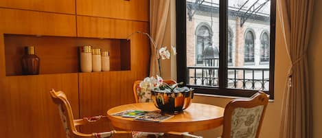 A dining area with a wooden table, floral chairs, and a large window with a view of buildings. Shelves with decorative items and a hanging light fixture are also visible.