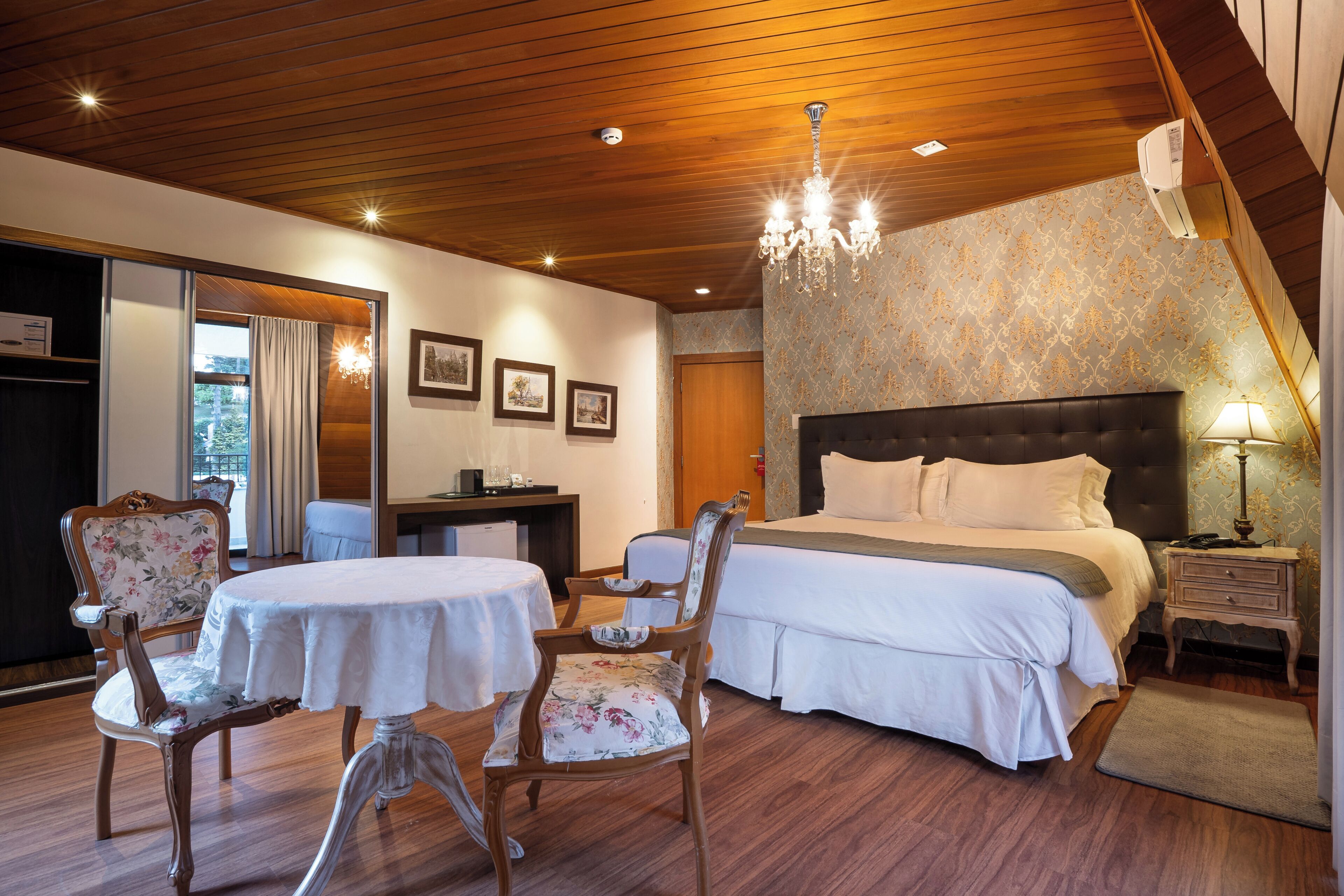 Quarto de hotel com uma cama, duas cadeiras, uma mesa, escrivaninha, abajur e lustre.