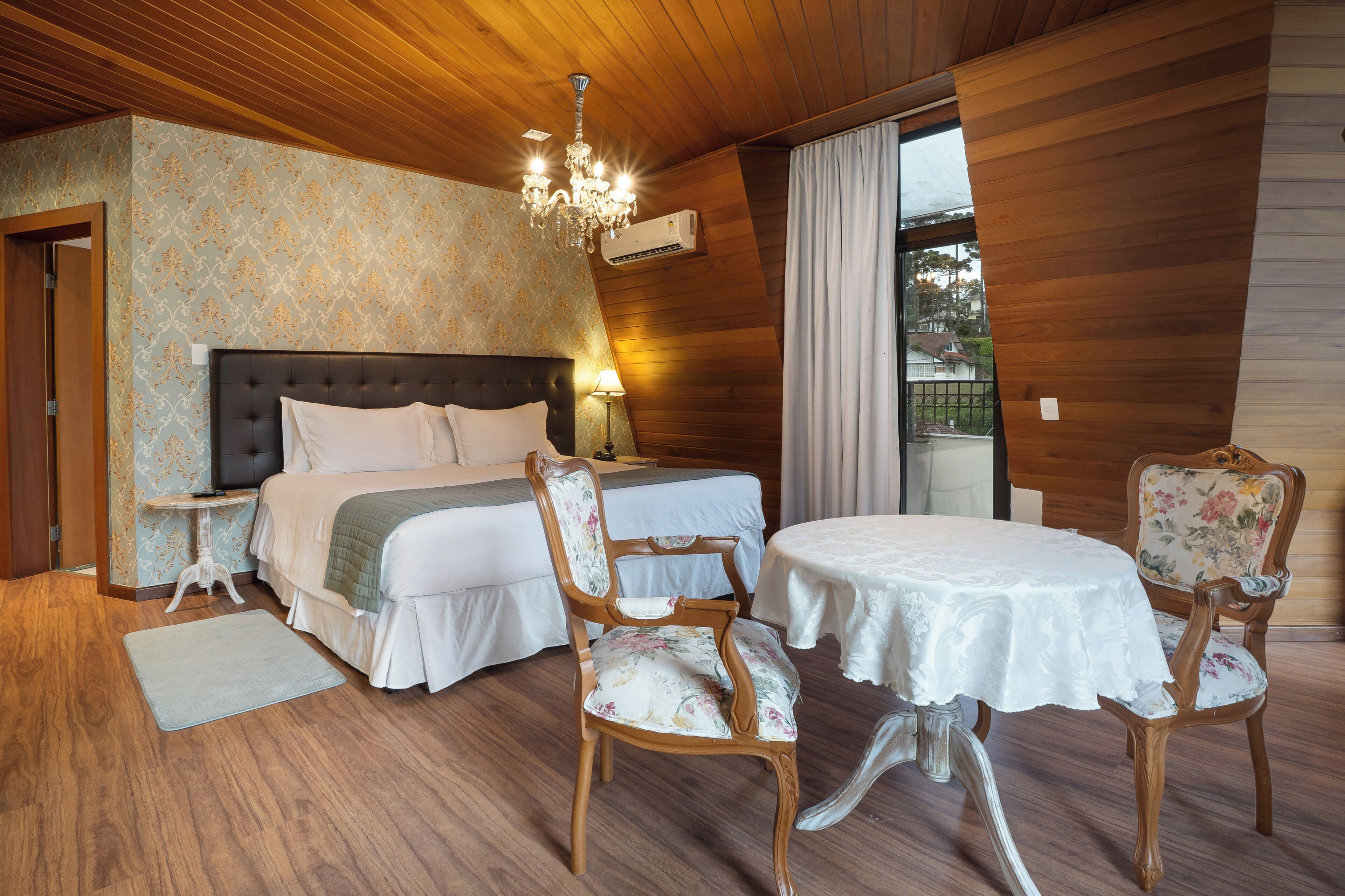 A bedroom with a large bed, two chairs, a table, a chandelier, and a view of the outdoors.