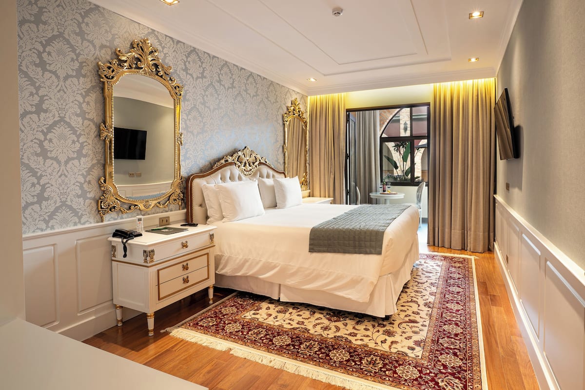 Un dormitorio con un espejo grande y ornamentado, una cama con ropa de cama blanca, una mesita de noche con lámpara y una alfombra estampada.