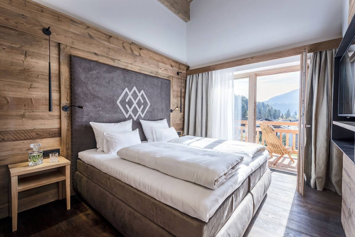 Un dormitorio con una cama grande, cabecera de madera, una mesita de noche y vistas a un balcón con una silla.