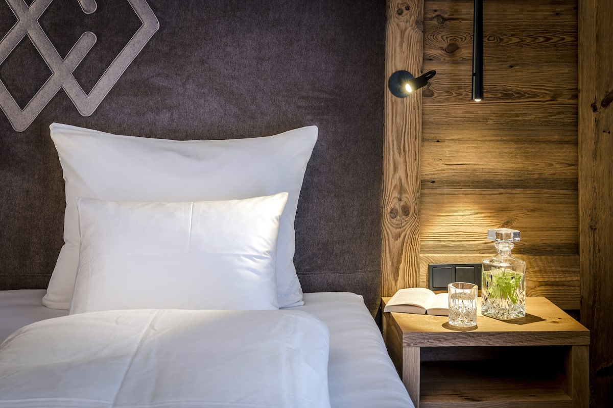 Una cama bien hecha con sábanas y almohadas blancas, una mesita de noche con un libro y un vaso de agua, y una pared de madera con un adorno metálico geométrico.