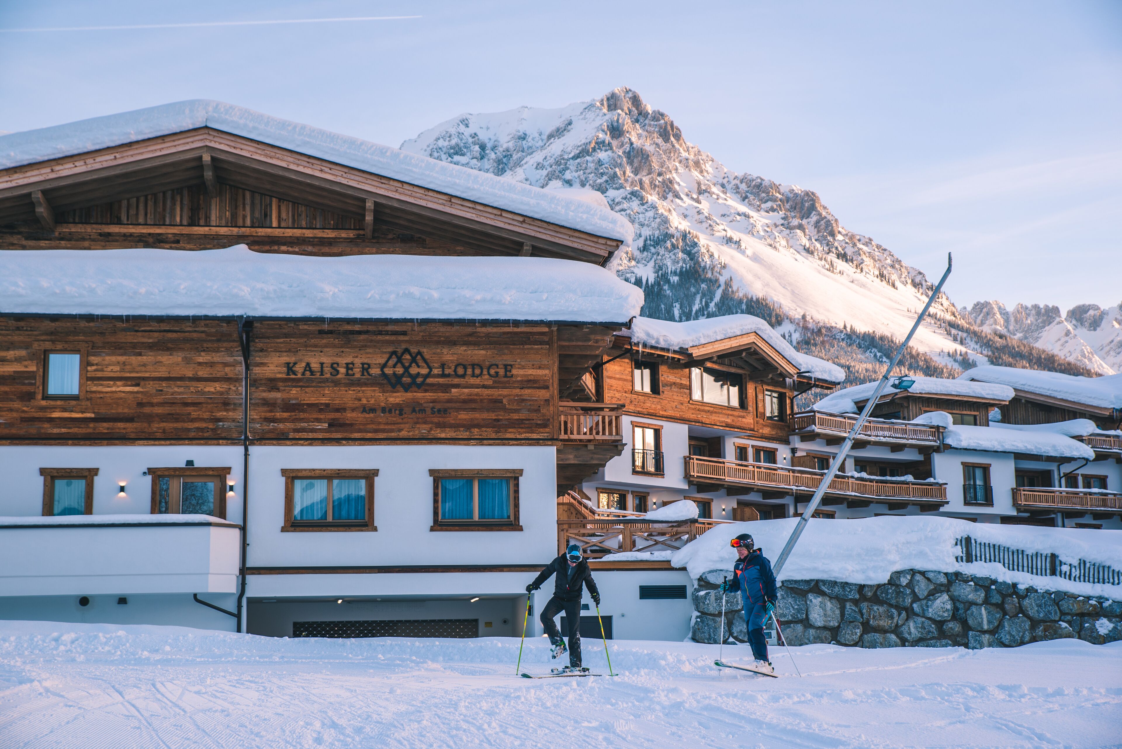 Een houten gebouw met de aanduiding "KAISER LODGE", skipistes en besneeuwde bergen op de achtergrond.