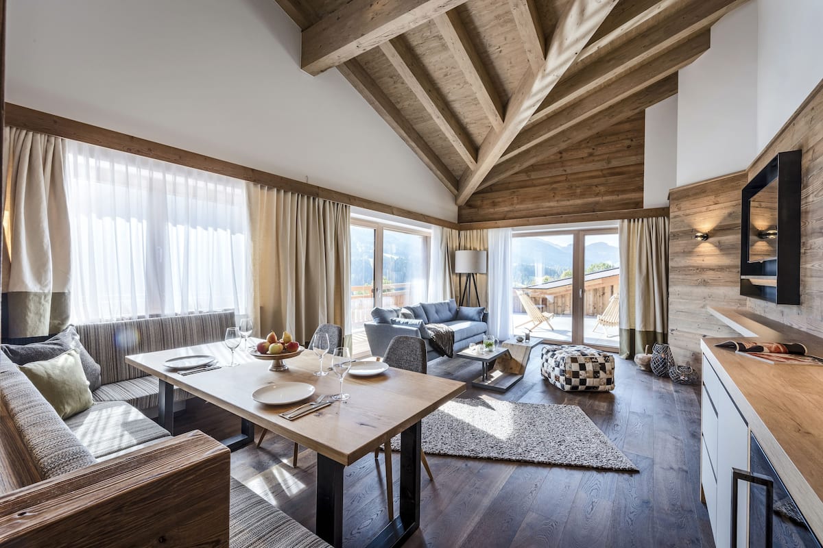 Amplia sala de estar con techo de madera, una mesa de comedor preparada para cuatro personas, un sofá y vistas a montañas a través de grandes ventanales.