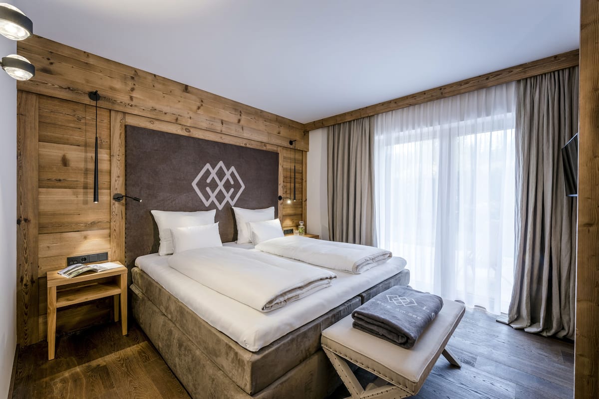 Una cama doble con ropa de cama blanca, un cabecero de madera, una mesita de noche con una revista y un ventanal con cortinas.